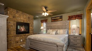 A bedroom with a stone fireplace, a bed with a headboard, a ceiling fan, and two bedside lamps.
