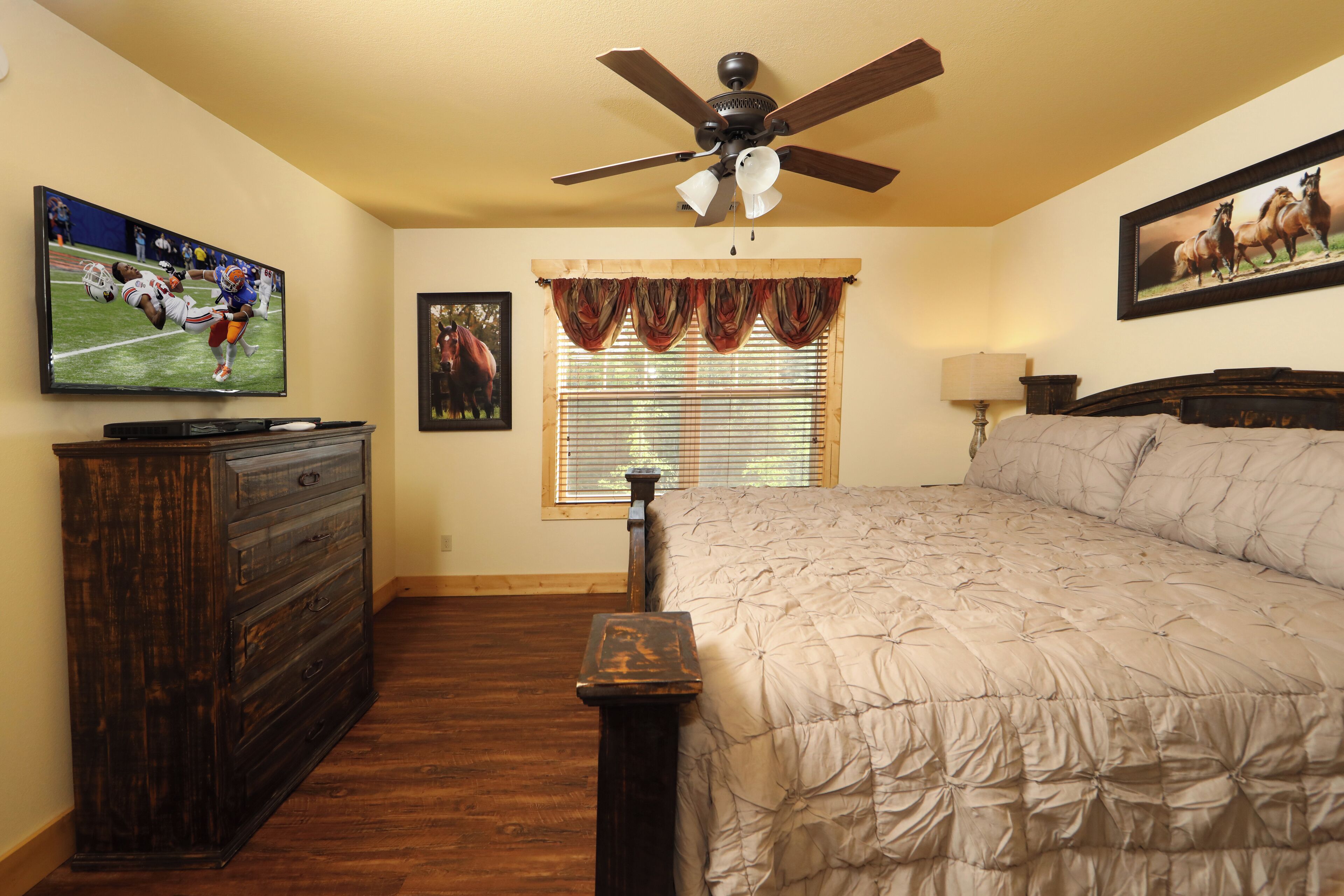 A bedroom with a bed, a ceiling fan, a dresser, a window with curtains, and a TV on the dresser.