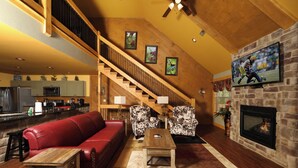 Ein geräumiges Wohnzimmer mit einem roten Ledersofa, zwei gemusterten Sessel, einem Holztisch und einem Kamin. Eine Treppe führt zu einer oberen Ebene, und ein Flachbildfernseher ist an der Wand befestigt.