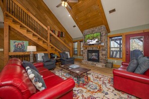 A cozy living room with a red leather sofa, a stone fireplace, a wooden staircase, and a television mounted on the wall.