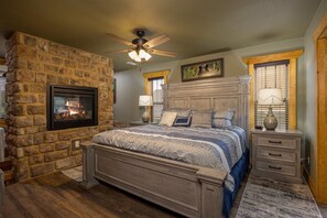 A bedroom with a stone fireplace, a bed with a wooden headboard, a nightstand with lamps, and a ceiling fan. - Mountain Palace - Indoor Pool, Theater, Game Room, Hot Tub, fire place (Gatlinburg)