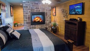 A bedroom with a stone fireplace, a bed with striped bedding, a ceiling fan, and a television on the wall. - Serene Oasis | Luxe 4BR Chalet w/ Indoor Pool, Theater, Game Room & Hot Tub (Gatlinburg)