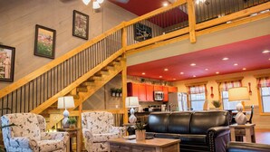 A spacious living area with a wooden staircase, a black leather sofa, patterned armchairs, and a dining table. - Serene Oasis | Luxe 4BR Chalet w/ Indoor Pool, Theater, Game Room & Hot Tub (Gatlinburg)