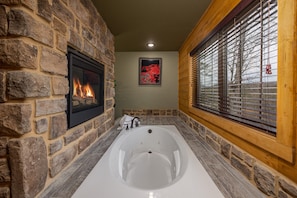 A bathroom with a stone wall, a fireplace, a large bathtub, and a window with blinds. - Serenity Peak -Lux Chalet, Indoor Saltwater Pool, Sauna, Theater & Game Loft (Gatlinburg)