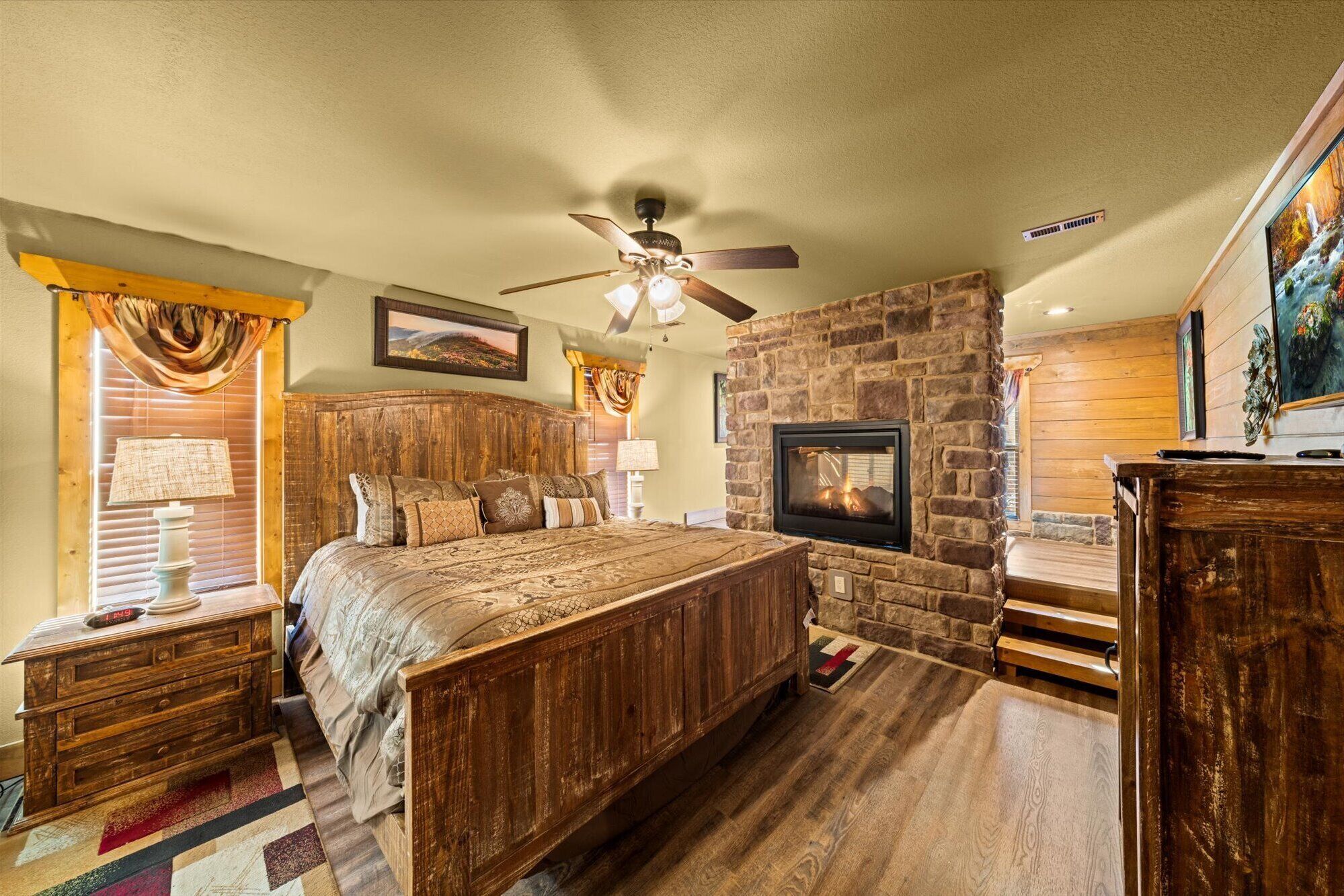 A bedroom with a wooden bed, a stone fireplace, a ceiling fan, and a painting on the wall.