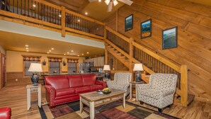 A spacious living room with a red sofa, patterned armchairs, and a wooden coffee table. A staircase with wooden railings leads to an upper level.