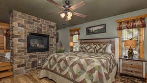 A bedroom with a stone fireplace, a bed with a quilt, a wooden nightstand, and a ceiling fan.