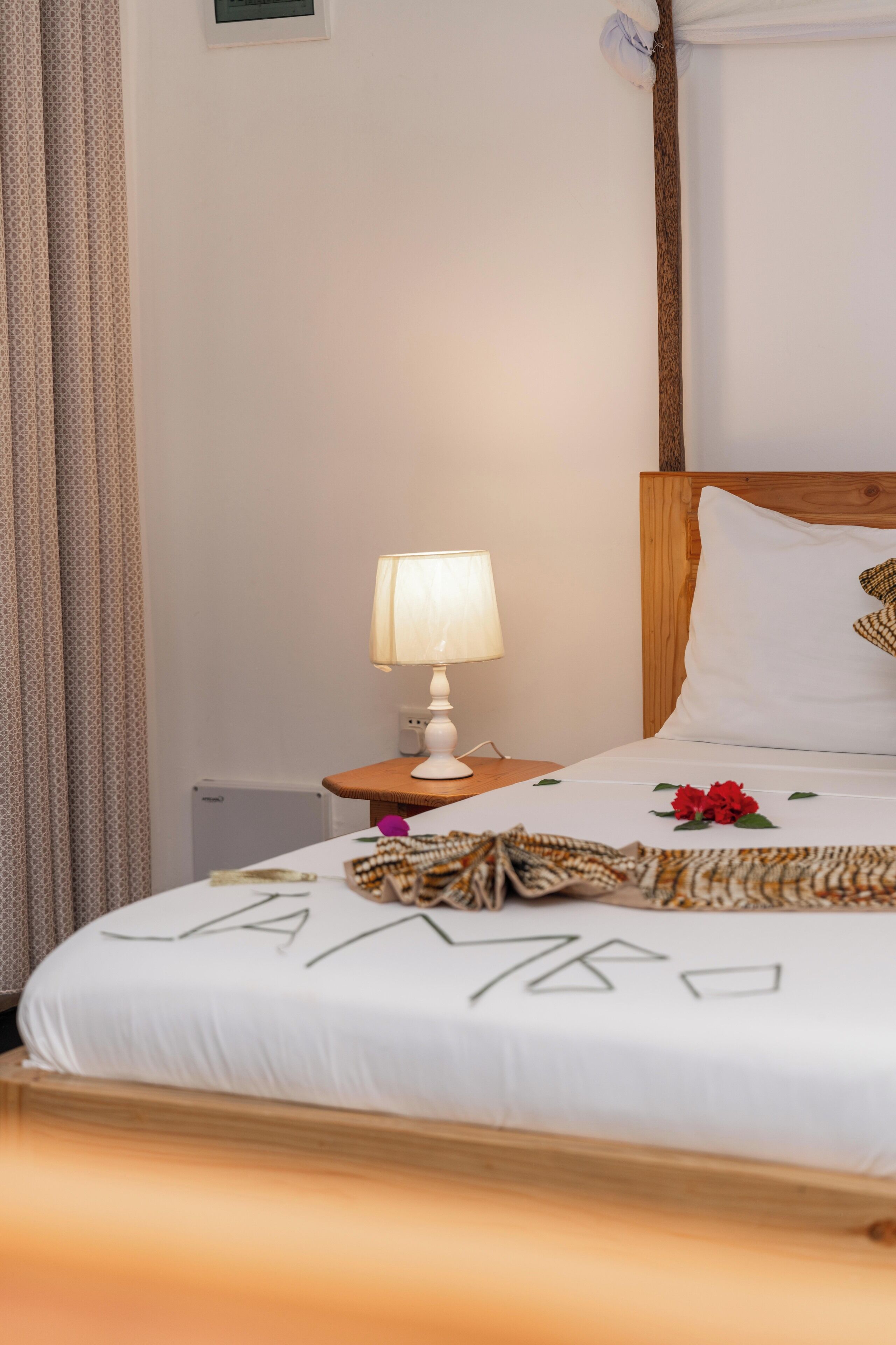 A neatly made bed with a white sheet and a decorative pillow, a bedside table with a lamp, and a curtain on the window.