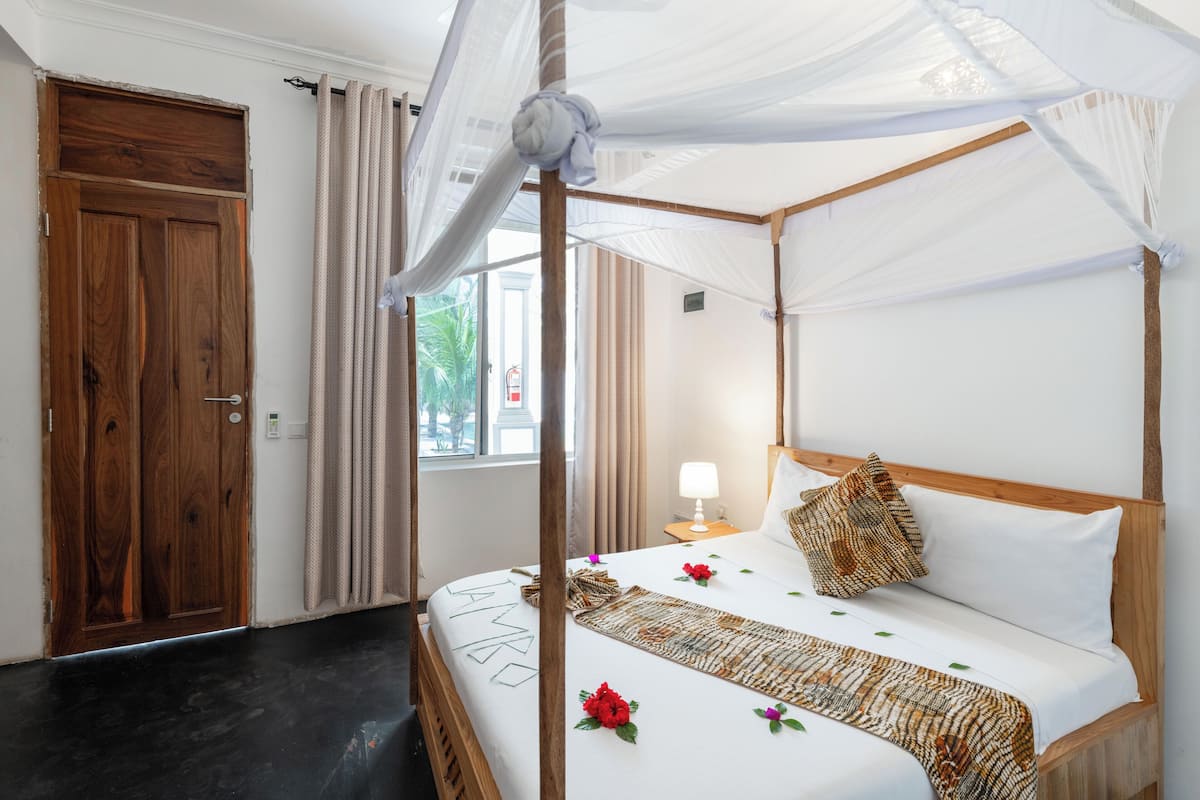 A four-poster bed with a canopy, a wooden door, a window with curtains, and a bedside table with a lamp.