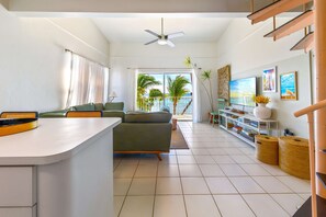 A modern living room with a sofa, a TV, and a balcony with a view of palm trees. - Moon Bay 2 Bdr Beachfront Condo with Pool and Tennis (Bodden Town)