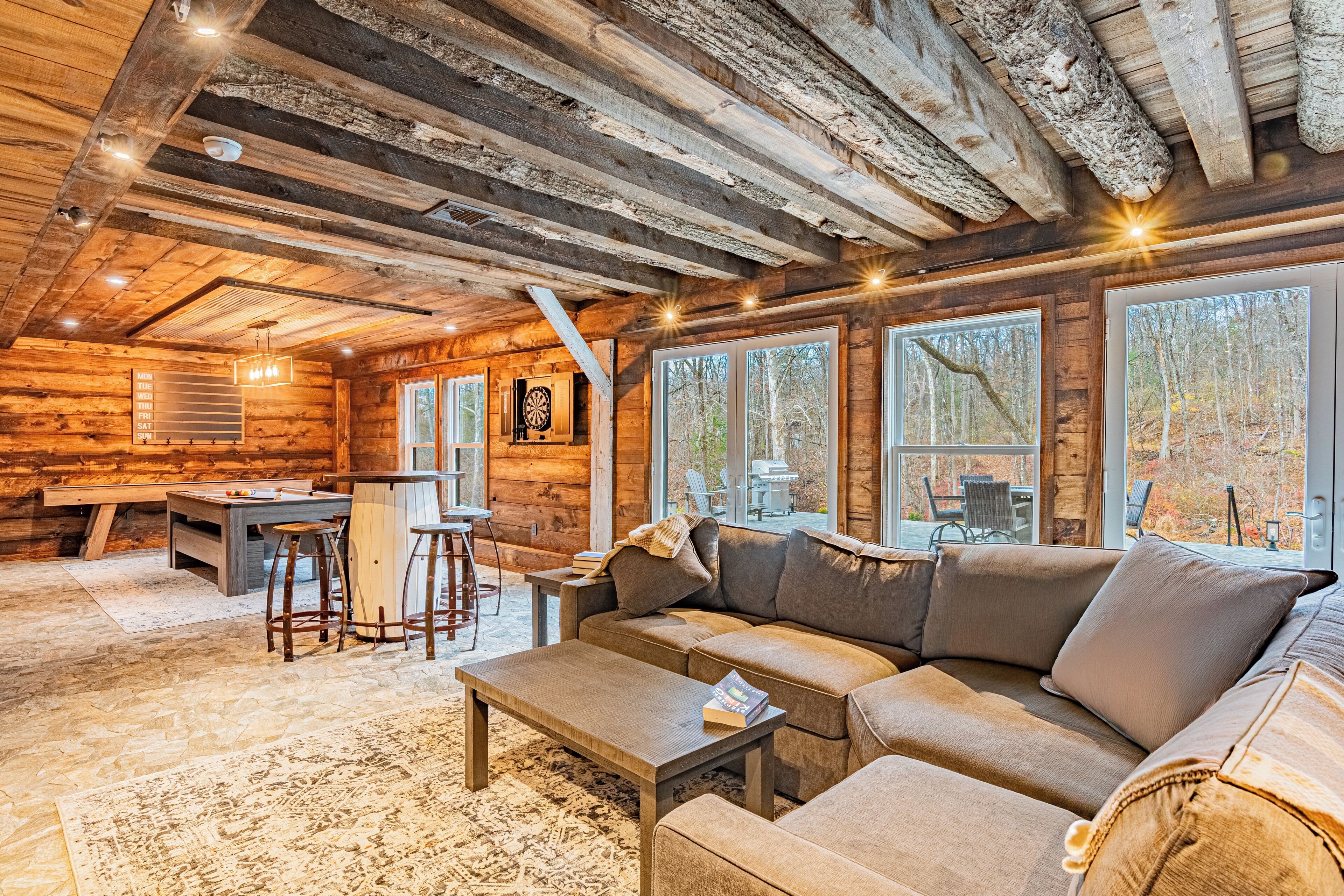 A cozy cabin interior with a wooden ceiling, a large sofa, a dining table, and a bar area.