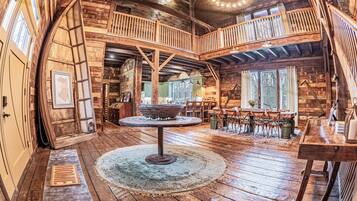A rustic cabin interior with a central wooden table, a balcony, and a dining area.
