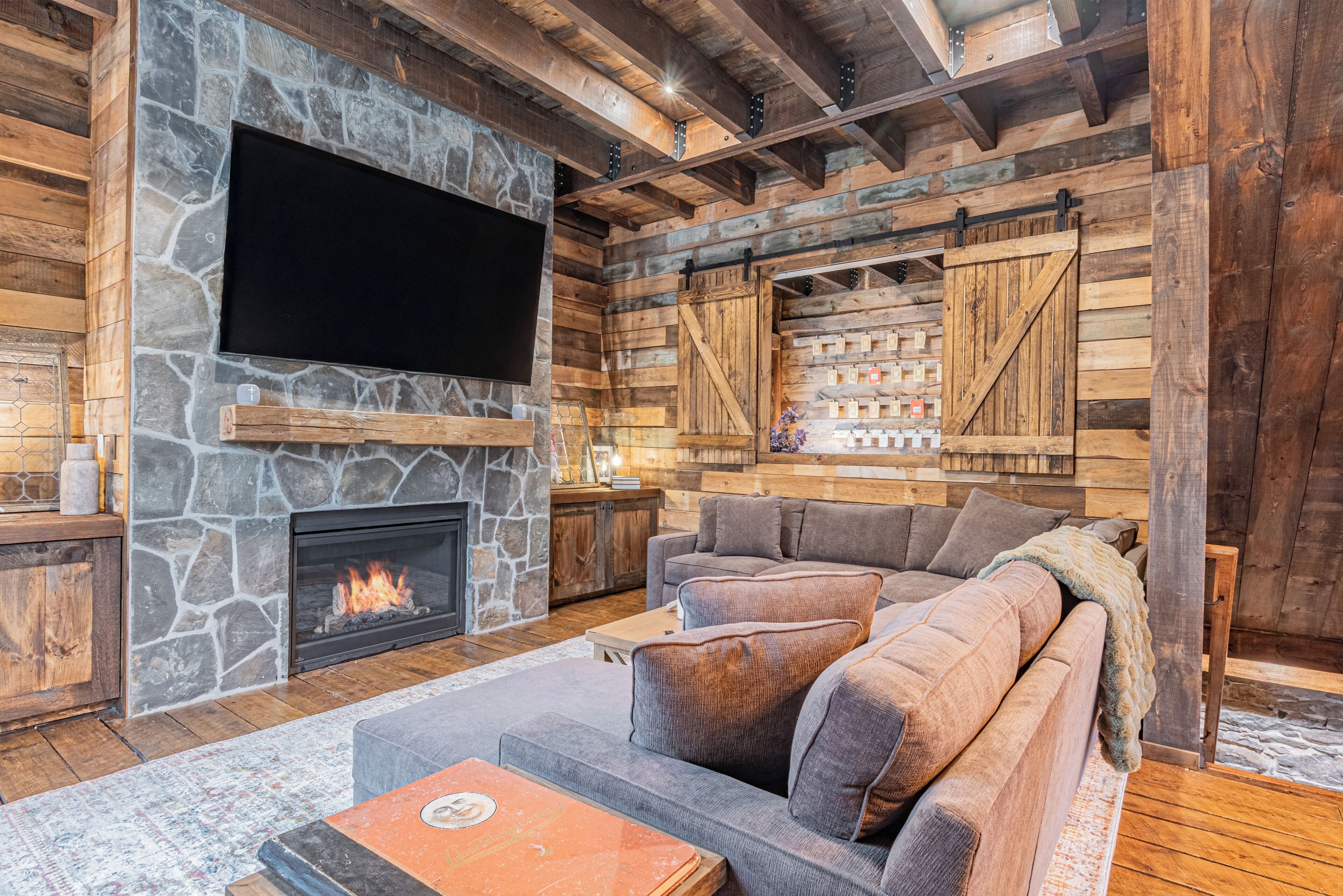 A cozy living room with a stone fireplace, a large flat-screen TV, and a wooden ceiling.