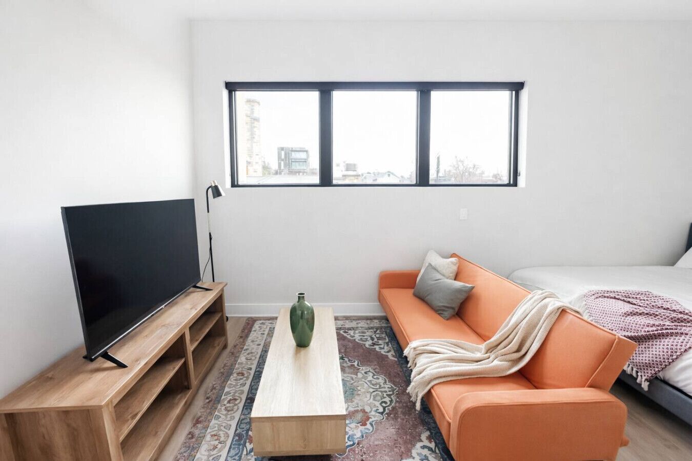 A modern living room with a flat-screen TV on a wooden stand, a green vase on a coffee table, an orange sofa with cushions, and a large window with a view of buildings.