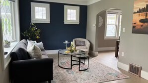 A living room with a dark blue accent wall, two windows, a sofa, a chair, a coffee table, and a rug.