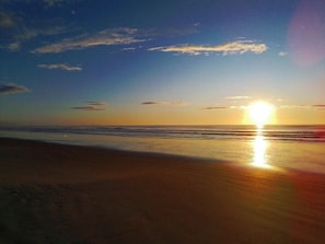 Ein Strand mit untergehendem Sonnenuntergang am Horizont, dessen Licht sich im Wasser spiegelt.