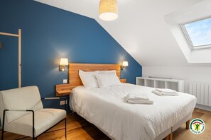 A bedroom with a sloped ceiling, a bed with white linens, a chair, and two wall-mounted lamps. - Gîtes de France® - La Grange (BIVILLE LA BAIGNARDE)