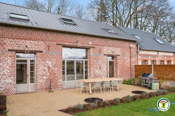 A brick building with large windows and a patio area with a table and chairs. - Gîtes de France® - La Grange (BIVILLE LA BAIGNARDE)