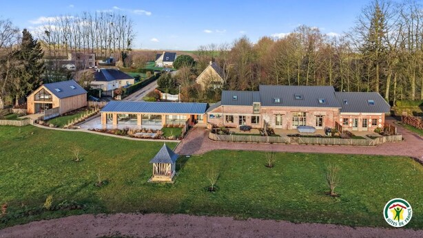 Un vaste domaine comprenant plusieurs bâtiments, dont une maison principale et divers corps de ferme, le tout entouré d’une pelouse bien entretenue et d’arbres.