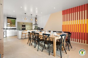 A modern kitchen with a dining area, featuring a large island, pendant lights, and a colorful wall. - Gîtes de France® - La Grange (BIVILLE LA BAIGNARDE)