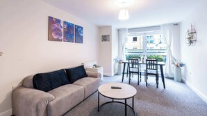 A living room with a sofa, a round coffee table, and a dining area with a table and chairs. There are two paintings on the wall, a ceiling light, and a window with curtains. - Spacious 2BR Flat - Bounds Green, Private Parking (London)