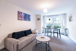 A living room with a sofa, a round coffee table, and a dining area with a table and chairs. There are two paintings on the wall, a ceiling light, and a window with curtains. - Spacious 2BR Flat - Bounds Green, Private Parking (London)