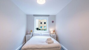 A hotel room with a large bed, two bedside tables, a window with a view of a building, and a small decorative item on the wall. - Spacious 2BR Flat - Bounds Green, Private Parking (London)