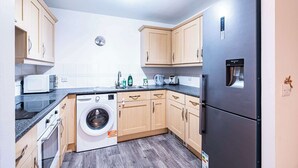 A compact kitchen with wooden cabinets, a washing machine, a refrigerator, and a microwave. - Spacious 2BR Flat - Bounds Green, Private Parking (London)