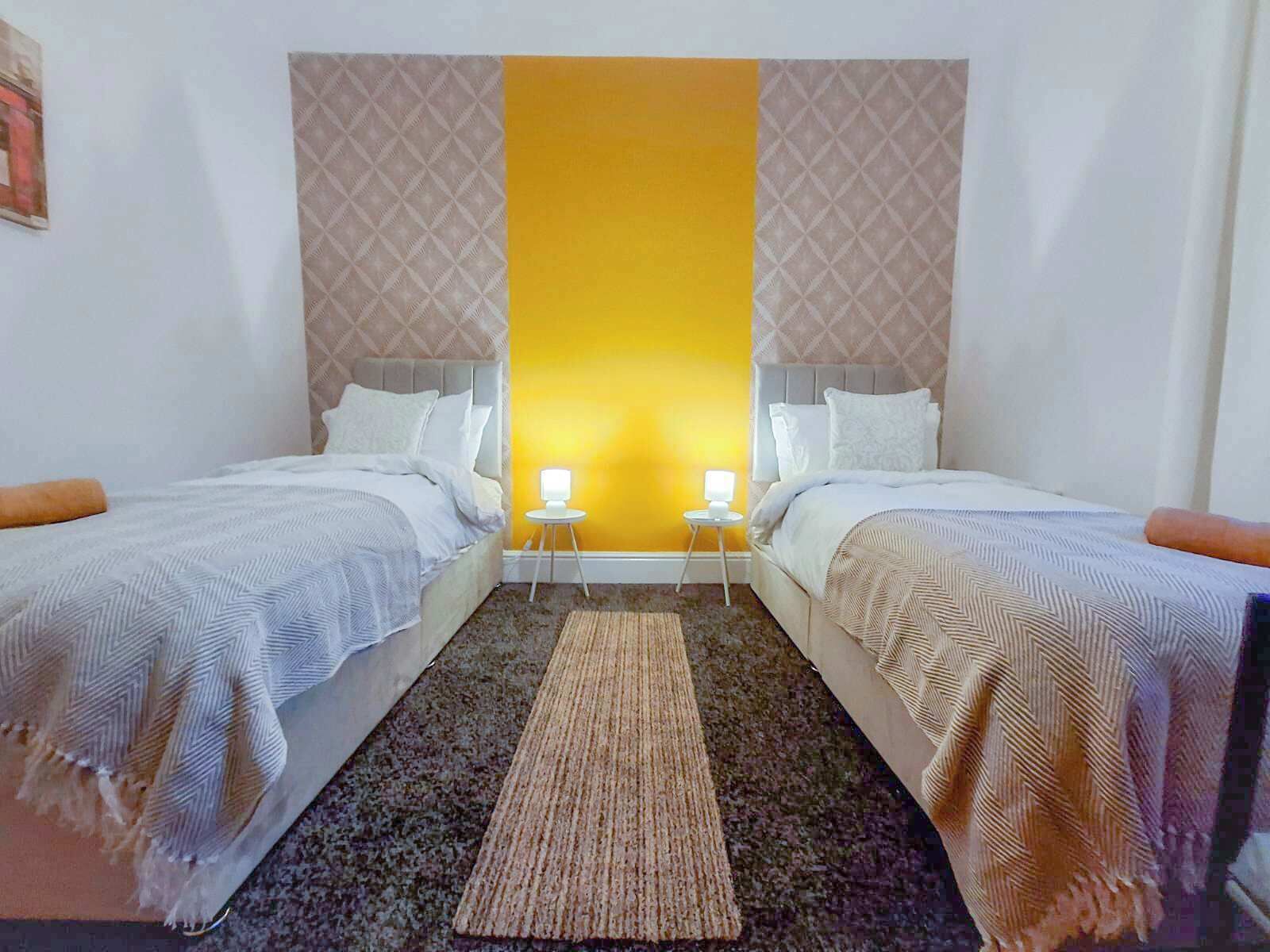 Een hotelkamer met twee bedden, een gele accentmuur in het midden en een kamerbreed tapijt met patroon.