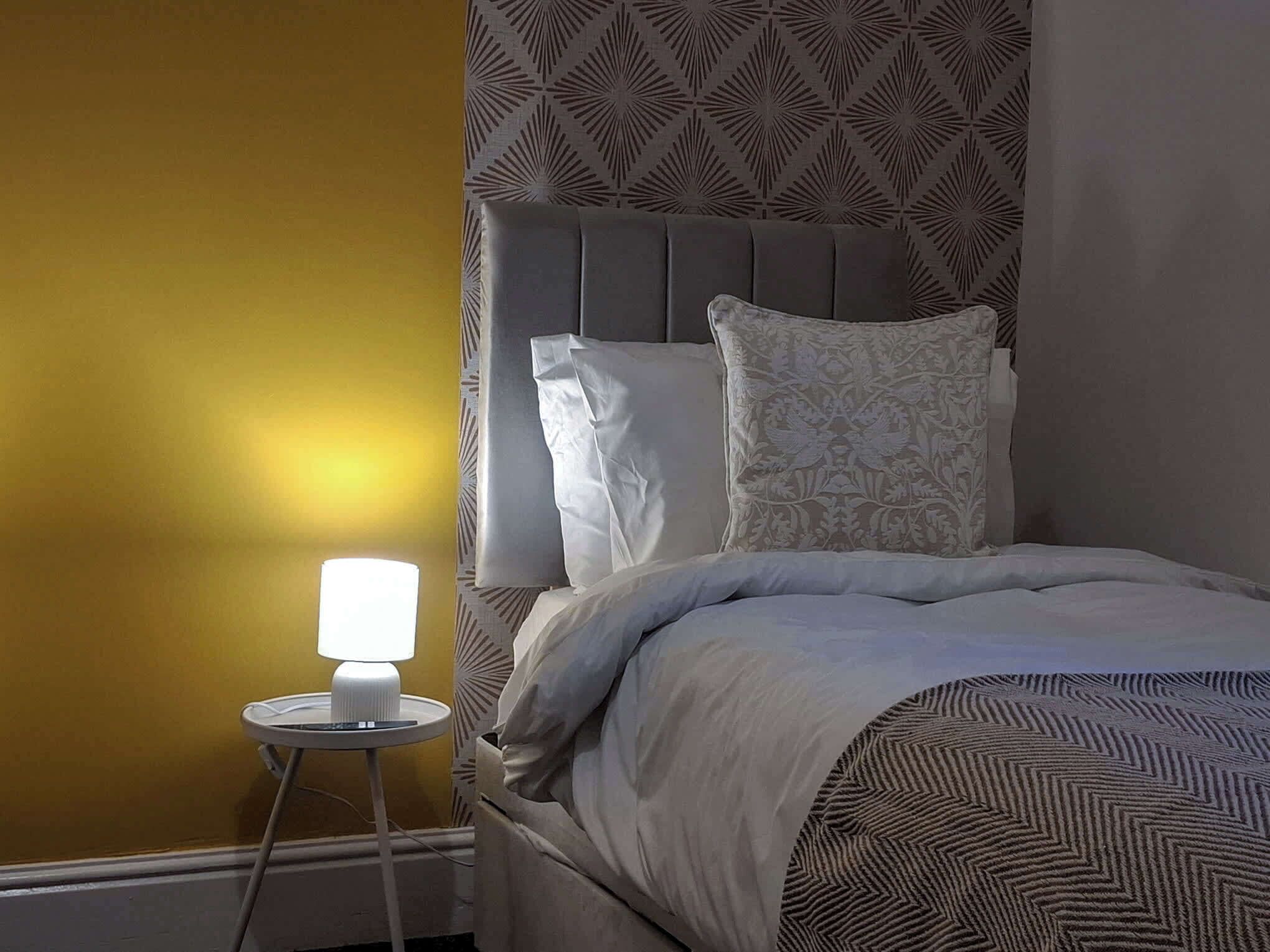 A neatly made bed with a patterned bedspread, a bedside lamp, and a patterned wall.