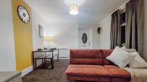 Een woonkamer met een bank, een eettafel met stoelen, een lamp, een klok aan de muur en een deur op de achtergrond.