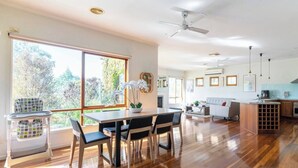 A spacious living area with a wooden floor, a dining table with chairs, a sofa, a kitchen area with a built-in island, and a large window with a view of trees. - Cascade Cottage: Spacious Family Retreat in Keysborough Garden Parking Sleeps 8 (Keysborough)