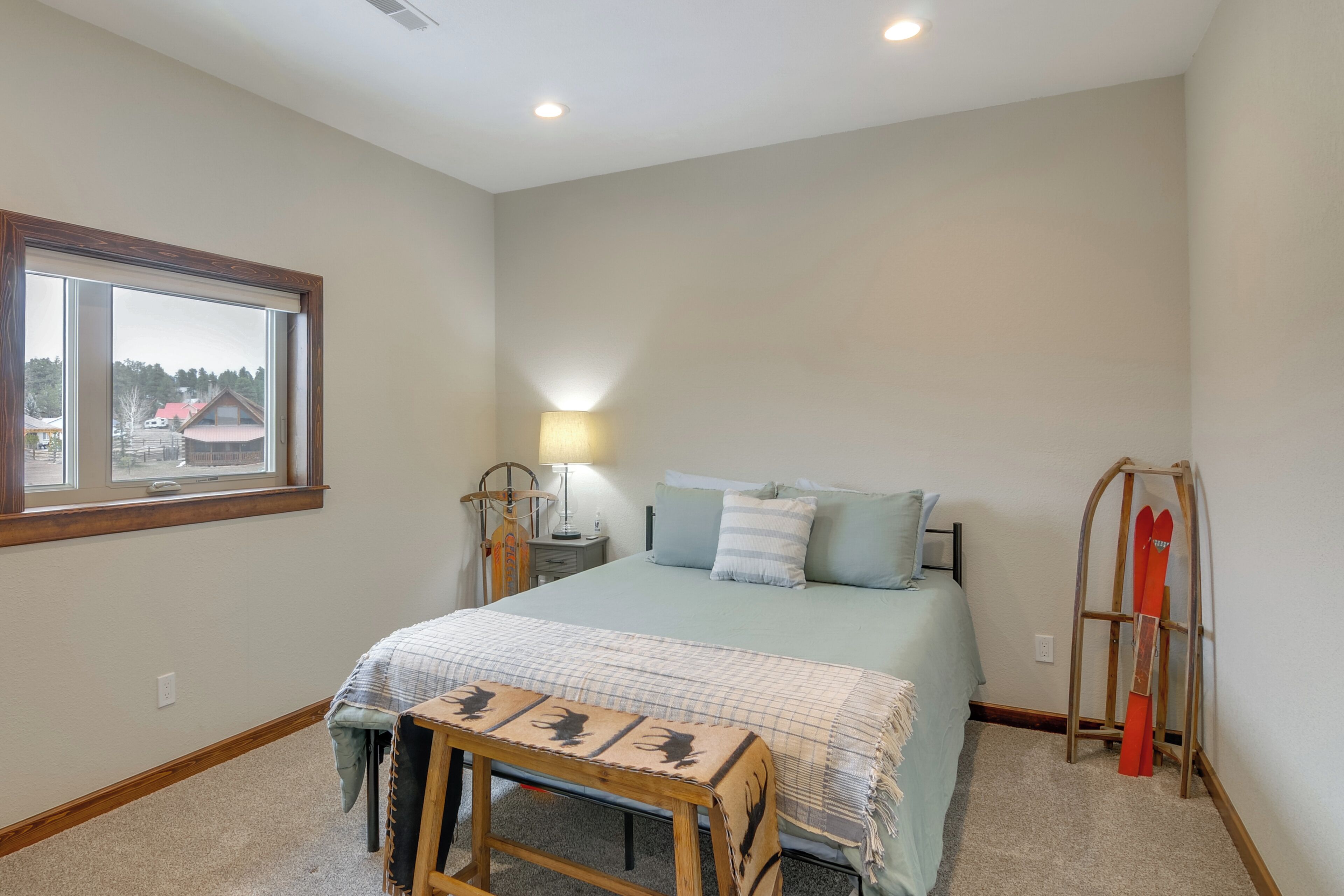 A bedroom with a bed, a bedside table, a lamp, a window, and a ski rack.