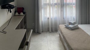 Ein Hotelzimmer mit Bett, Wandfernseher, Schreibtisch mit Computer und Fenster mit durchscheinenden Vorhängen.
