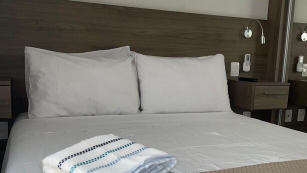 Ein ordentlich bezogenes Bett mit weißen Leinentüchern und einem Stapel gefalteter Handtücher auf einer strukturierten, beige farbenen Bettdecke.