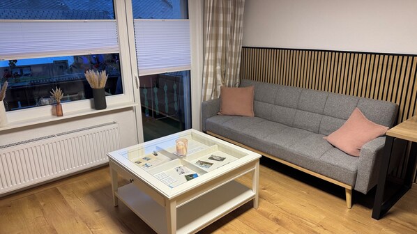 A modern living room with a grey sofa, a glass coffee table, and a window with blinds.