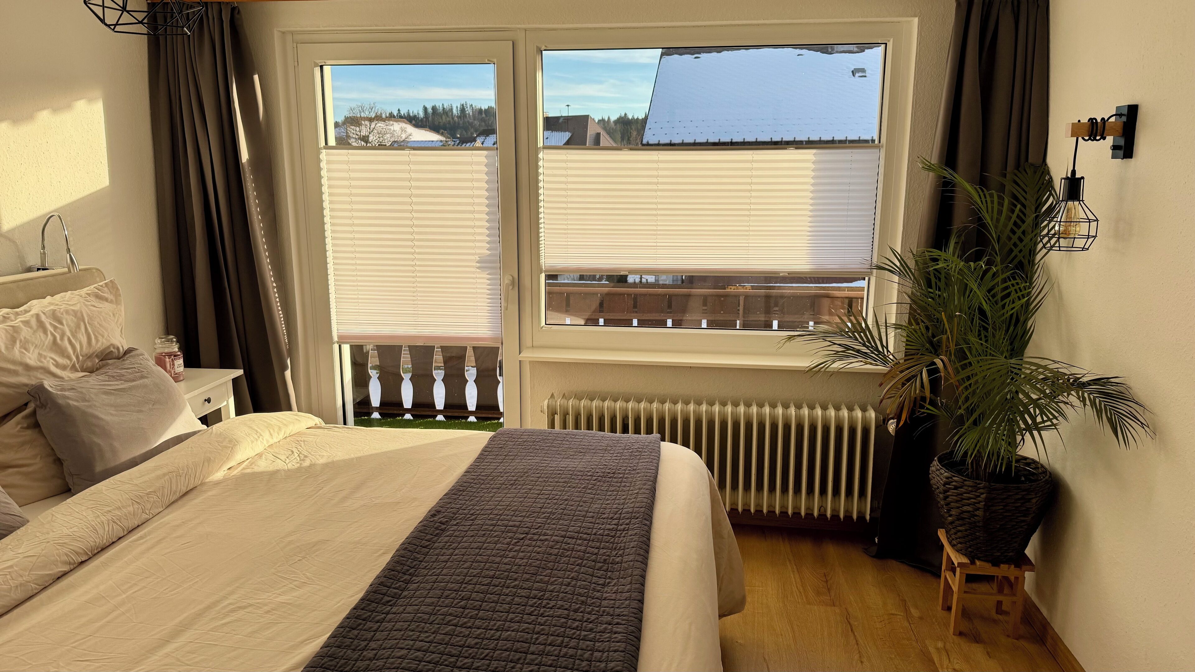 A bedroom with a bed, a radiator, a window with blinds, a potted plant, and a small stool.