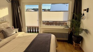 Ein Schlafzimmer mit einem Bett, einem Heizkörper, einem Fenster mit Jalousien, einer Topfpflanze und einem kleinen Hocker.