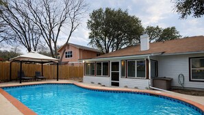 A backyard with a swimming pool, a covered patio with chairs, and a house with a covered entryway.