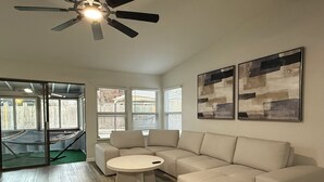A modern living room with a sectional sofa, a round coffee table, and a ceiling fan. There are two windows with blinds, and artwork on the wall.