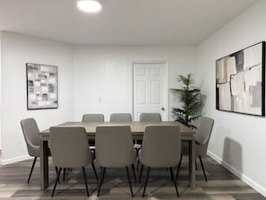 Uma sala de jantar com uma mesa retangular, seis cadeiras, uma planta em vaso e duas pinturas abstratas nas paredes.