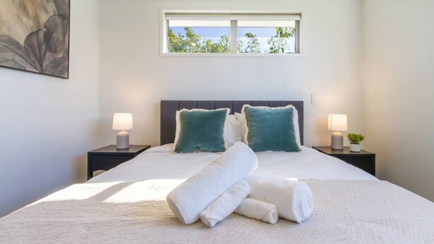 Una cama bien tendida con sábanas blancas y toallas enrolladas, dos almohadas color turquesa y dos lámparas de noche sobre mesitas.