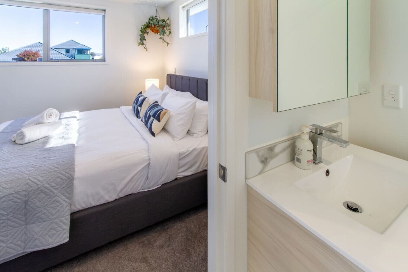 Une chambre à coucher comprenant un lit, des oreillers, une table de chevet et une plante suspendue, ainsi qu’une salle de bain avec un lavabo et un miroir.