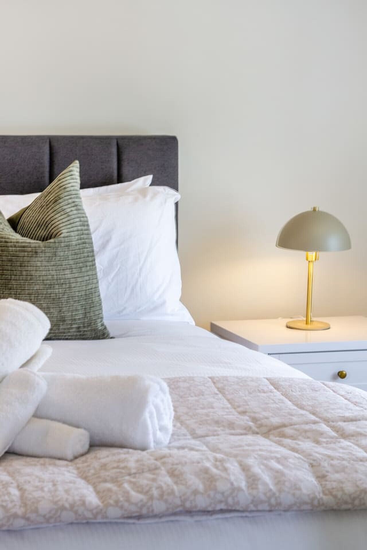 Een net opgemaakt bed met witte lakens, een grijs hoofdbord en een nachtkastje met een gouden lamp.