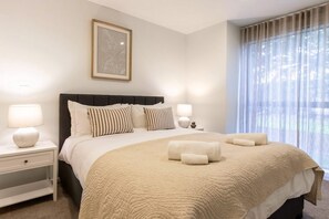 Quarto de hotel com uma cama grande, duas mesas de cabeceira, um criado-mudo, uma moldura com imagem na parede e uma janela com cortinas.