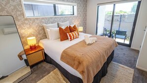A hotel room with a large bed, a nightstand with a lamp, a window, and a sliding glass door to an outdoor area.