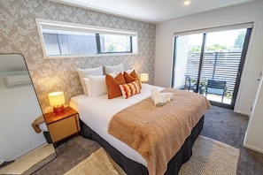 A hotel room with a large bed, a nightstand with a lamp, a window, and a sliding glass door to an outdoor area.