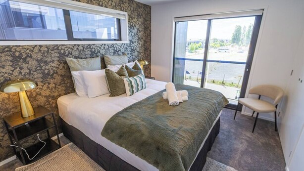 A bedroom with a large bed, a nightstand with a lamp, a chair, and a view of the outdoors through a large window.