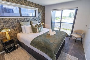 A bedroom with a large bed, a nightstand with a lamp, a chair, and a view of the outdoors through a large window.