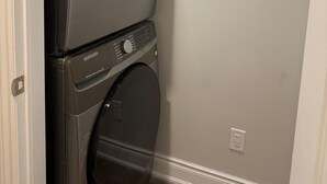 Un appareil combiné lave-linge et sèche-linge, installé en hauteur, dans une buanderie avec un mur sobre et une porte sur le côté gauche.